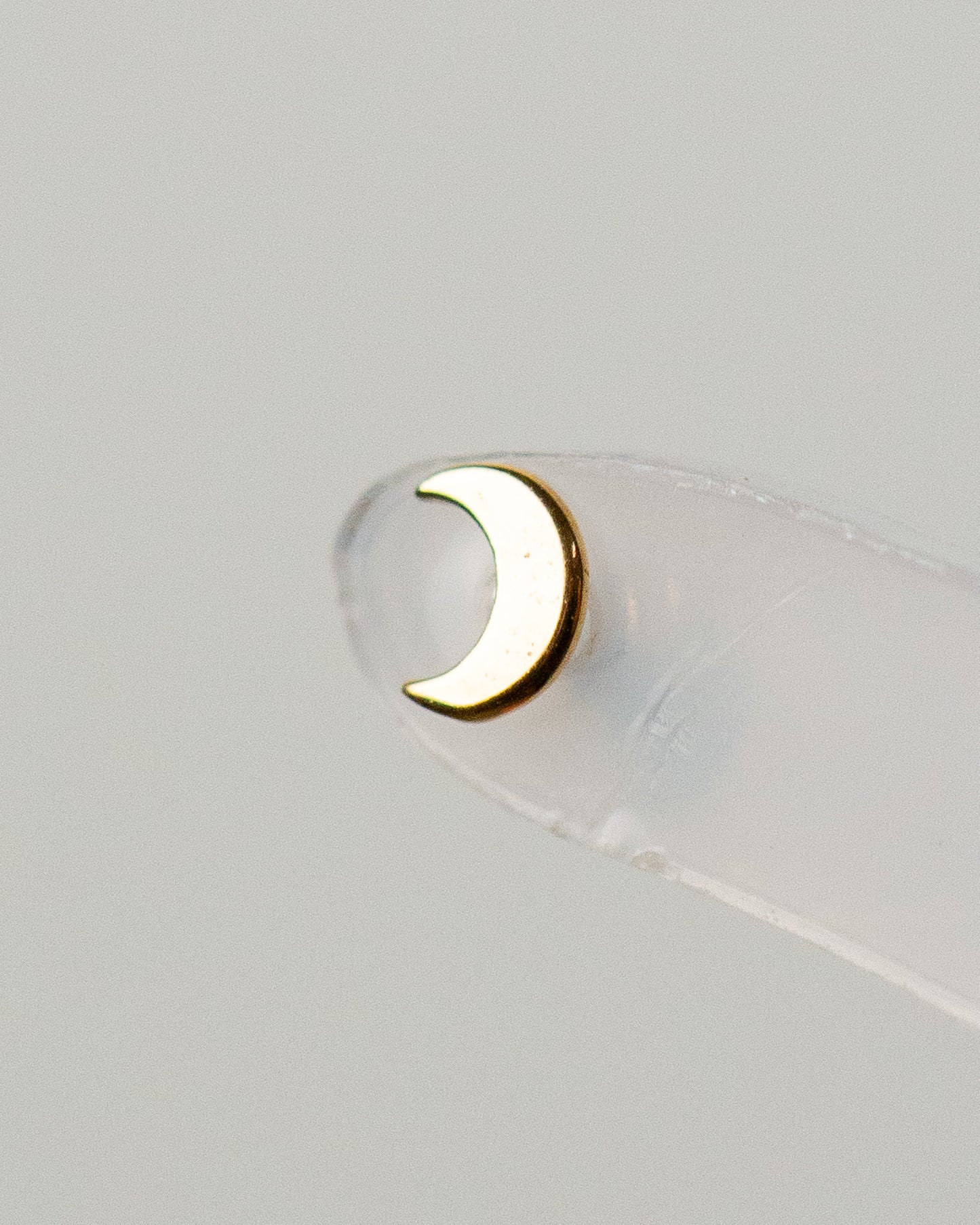 A close-up image of a 14K gold baby moon stud piercing jewelry on a clear backing.