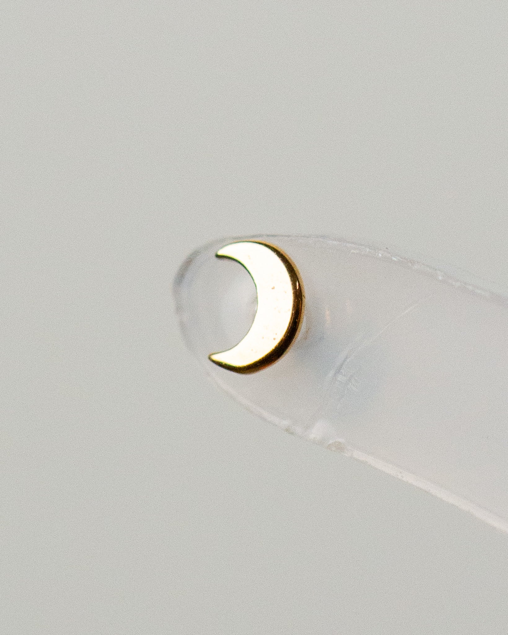 A close-up image of a 14K gold baby moon stud piercing jewelry on a clear backing.